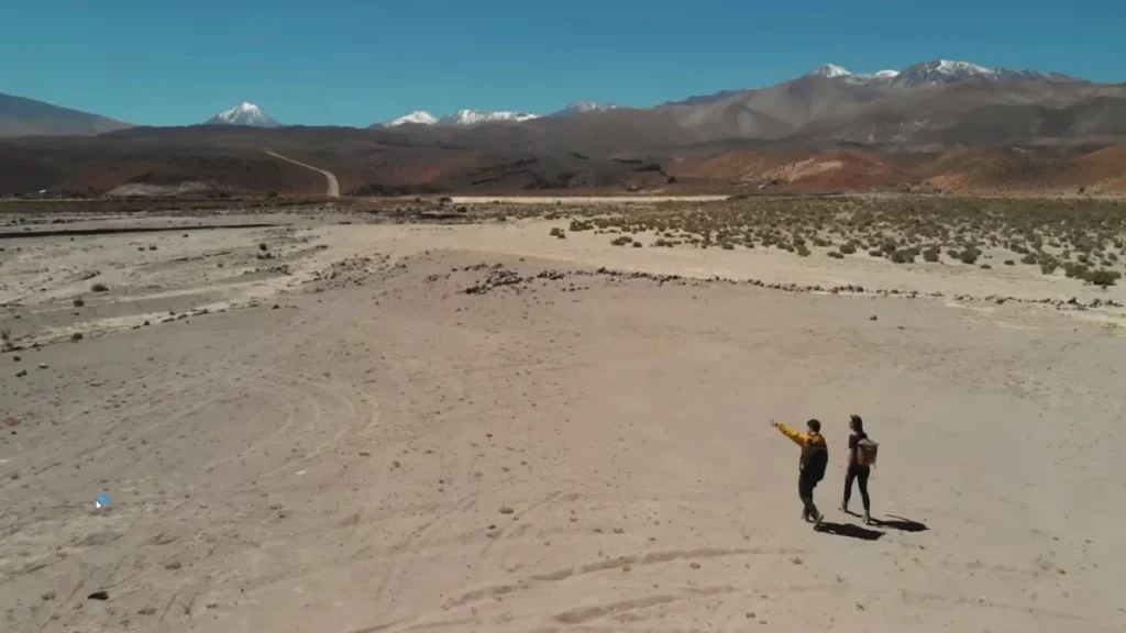 Duas pessoas caminhando no Deserto de Atacama com montanhas cobertas de neve ao fundo
