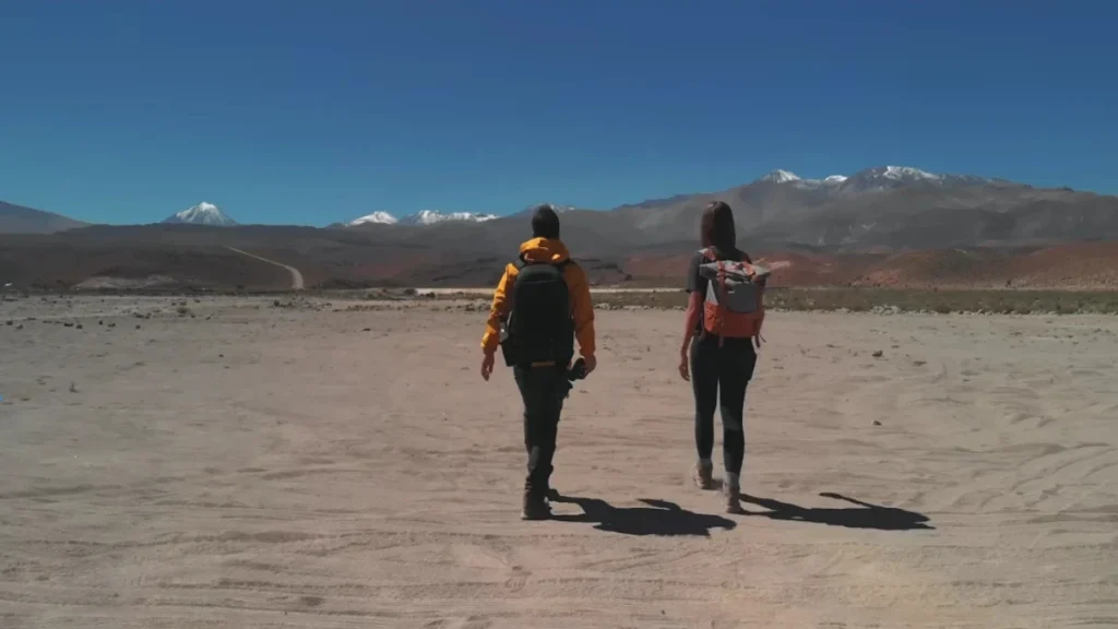 Dois viajantes explorando o Deserto de Atacama, com picos cobertos de neve ao fundo.