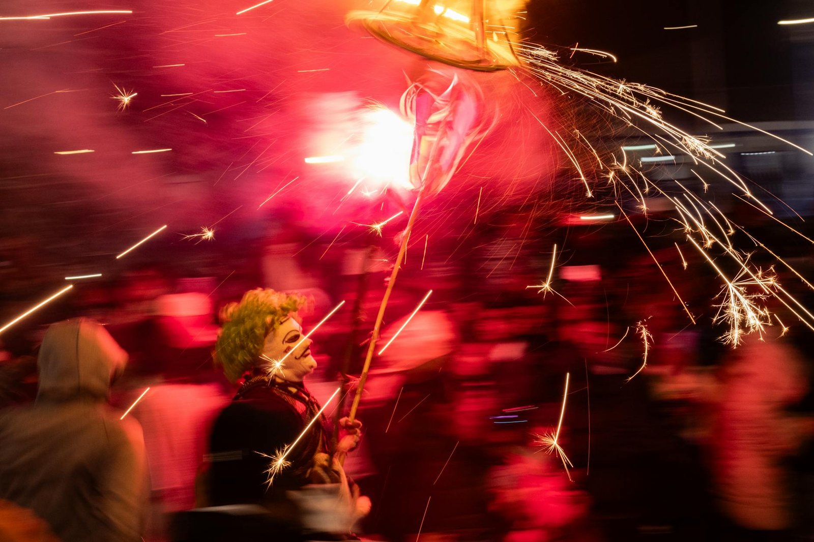 grupo de pessoas desfocadas com uma pessoa fantasiada segurando um graveto com pequenos fogos de artifício coloridos