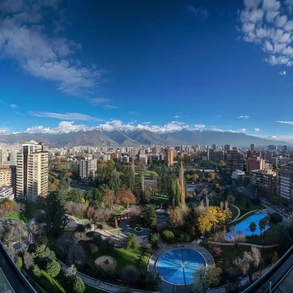 uma vista panorâmica de santiago em dezembro, com os andes majestosos ao fundo, céu azul radiante e um parque urbano vibrante em primeiro plano, refletindo a atmosfera calorosa e festiva do início do verão.