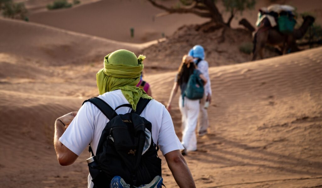 Grupo de viajantes caminhando em fila por dunas de areia em expedição de turismo de aventura.