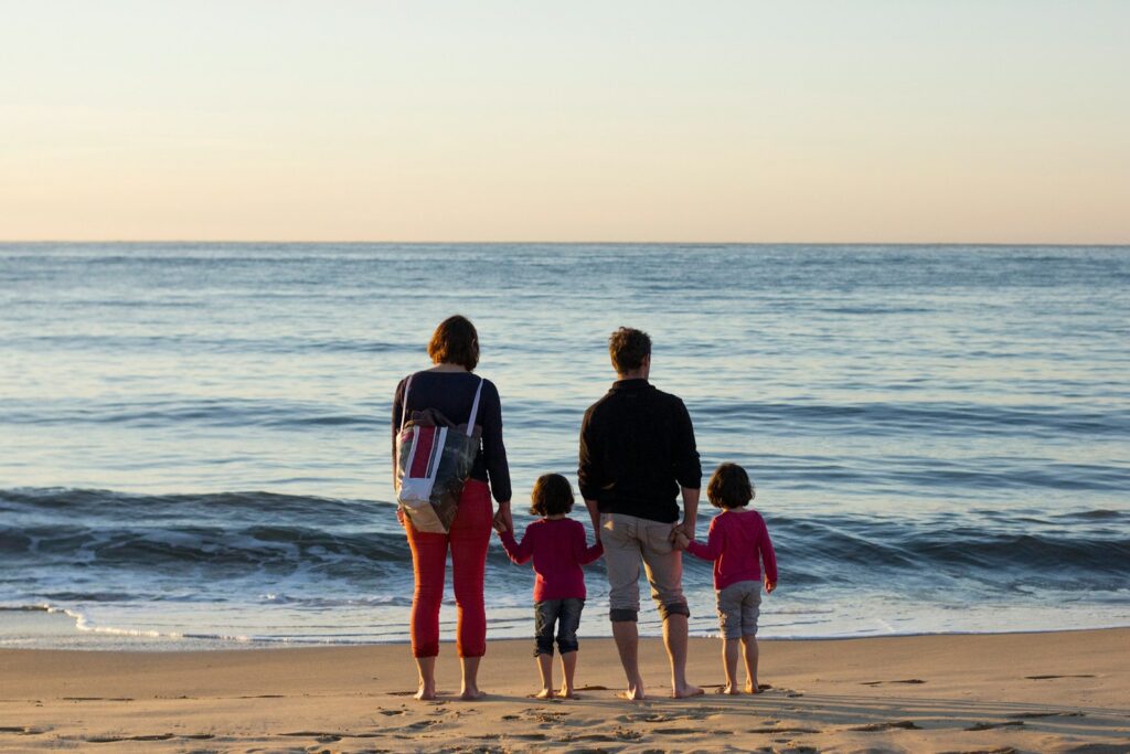 Família de quatro pessoas de costas observando o mar na praia durante o pôr do sol.
