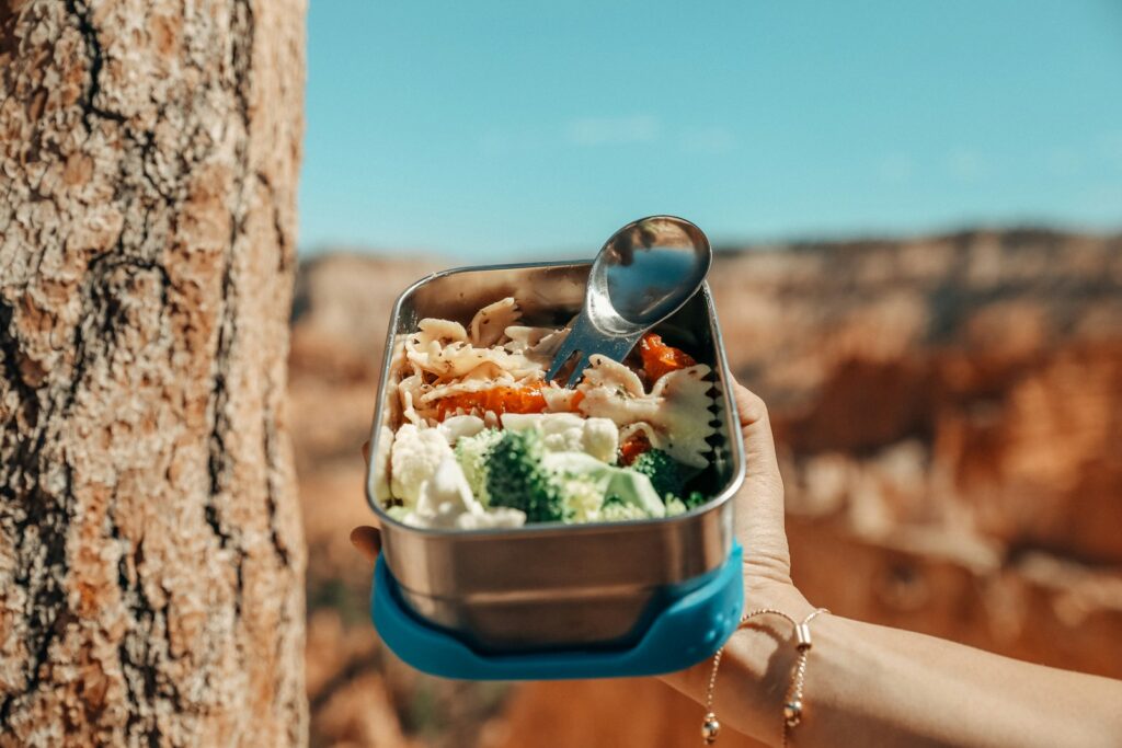 Mão segurando uma marmita térmica com macarrão e brócolis em um cenário de natureza ao ar livre.