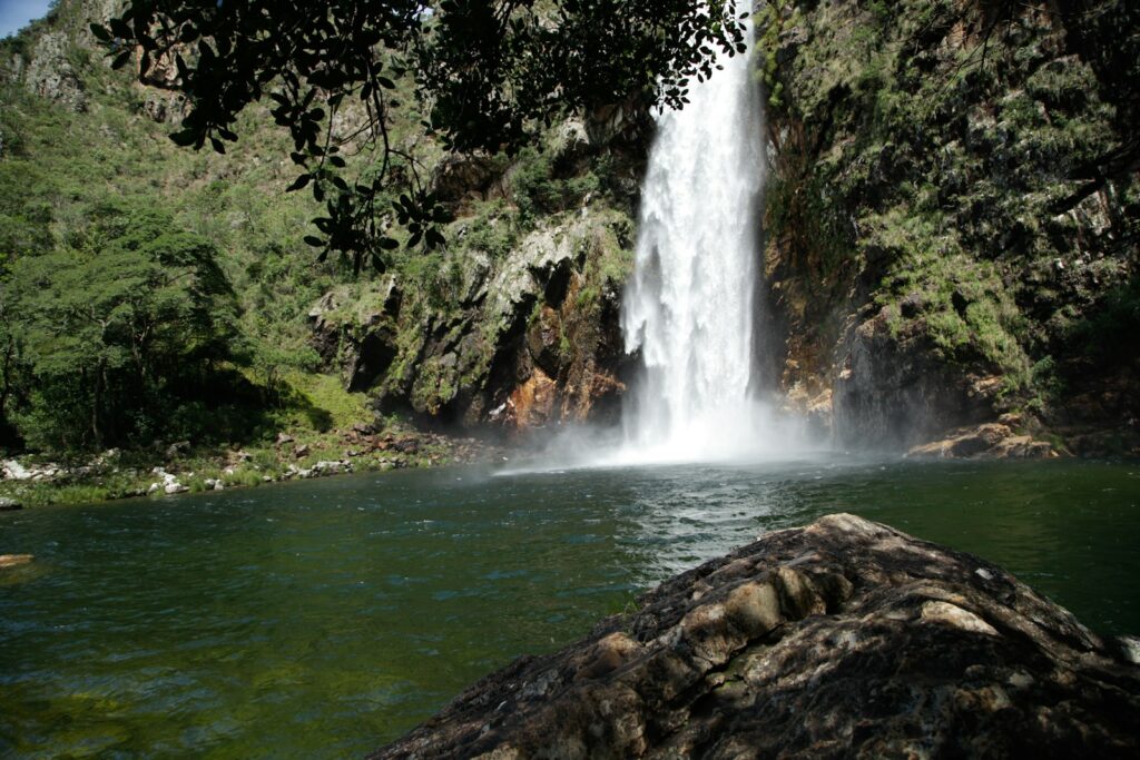 Queda d'água alta despencando de um paredão rochoso para um poço de águas verdes e cristalinas cercado por árvores.
