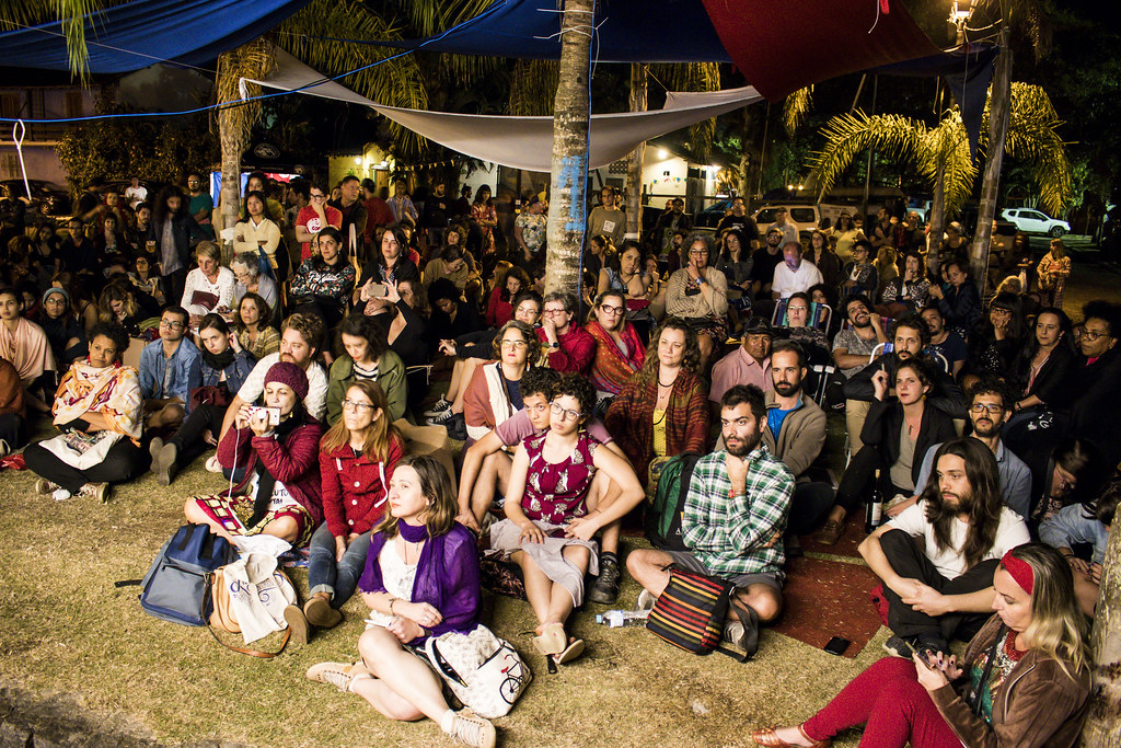 Multidão sentada na grama assistindo a uma palestra noturna ao ar livre na FLIP, sob tendas e iluminação cênica em Paraty.