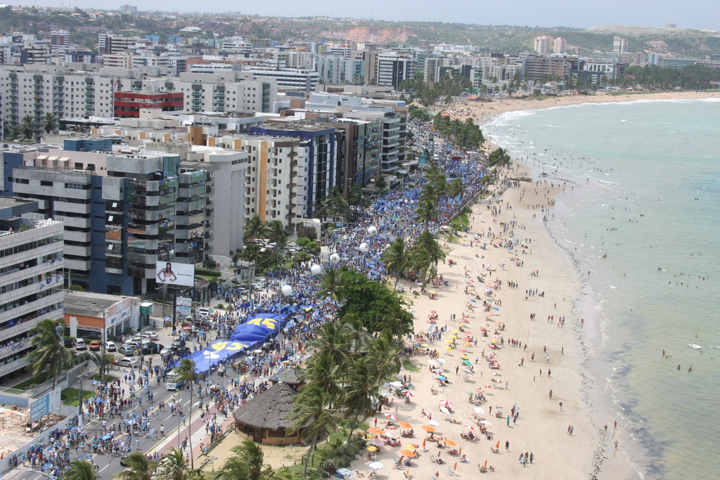 Vista aérea da orla de Maceió com calçadão movimentado, prédios modernos e mar verde-esmeralda ao fundo.