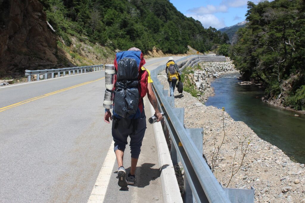 Dois viajantes com mochilas grandes caminhando pelo acostamento de uma estrada ao lado de um rio.