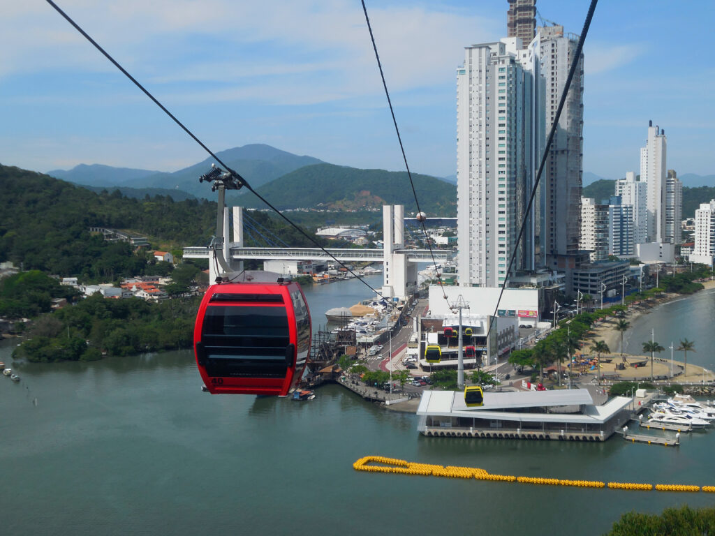 Bondinho vermelho do Parque Unipraias cruzando o rio com vista para os arranha-céus de Balneário Camboriú em um dia de sol.