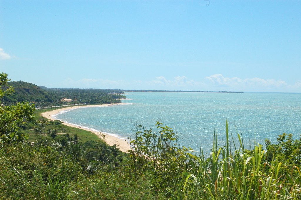 Vista do alto das falésias de Trancoso mostrando o mar azul turquesa e a longa faixa de areia cercada por vegetação nativa.