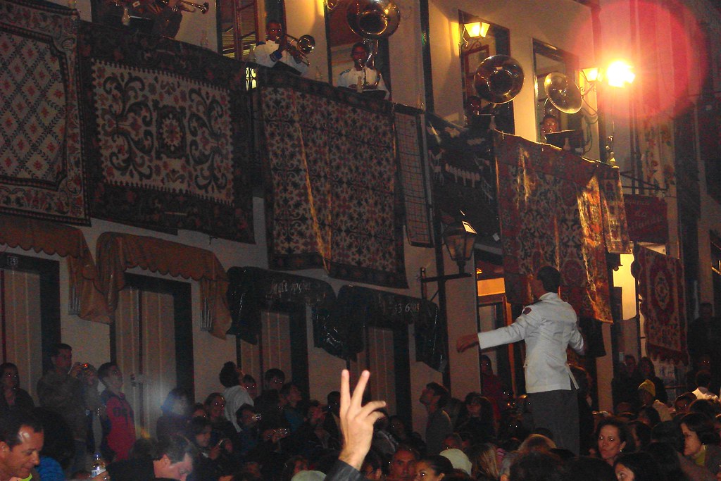 Músicos tocando nas sacadas de casarões coloniais decorados com tapetes durante a tradicional Vesperata em Diamantina, Minas Gerais, sob a regência de um maestro na rua.