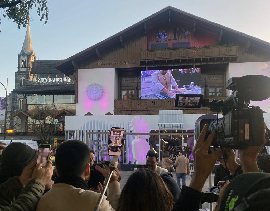 Multidão de fãs e jornalistas com celulares e câmeras erguidos, registrando a chegada de celebridades no tapete vermelho em frente ao Palácio dos Festivais, em Gramado.