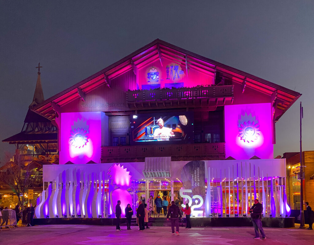 Fachada do Palácio dos Festivais iluminada em tons de rosa e roxo à noite, com o número 52 em destaque e um telão exibindo filmes durante o evento em Gramado.
