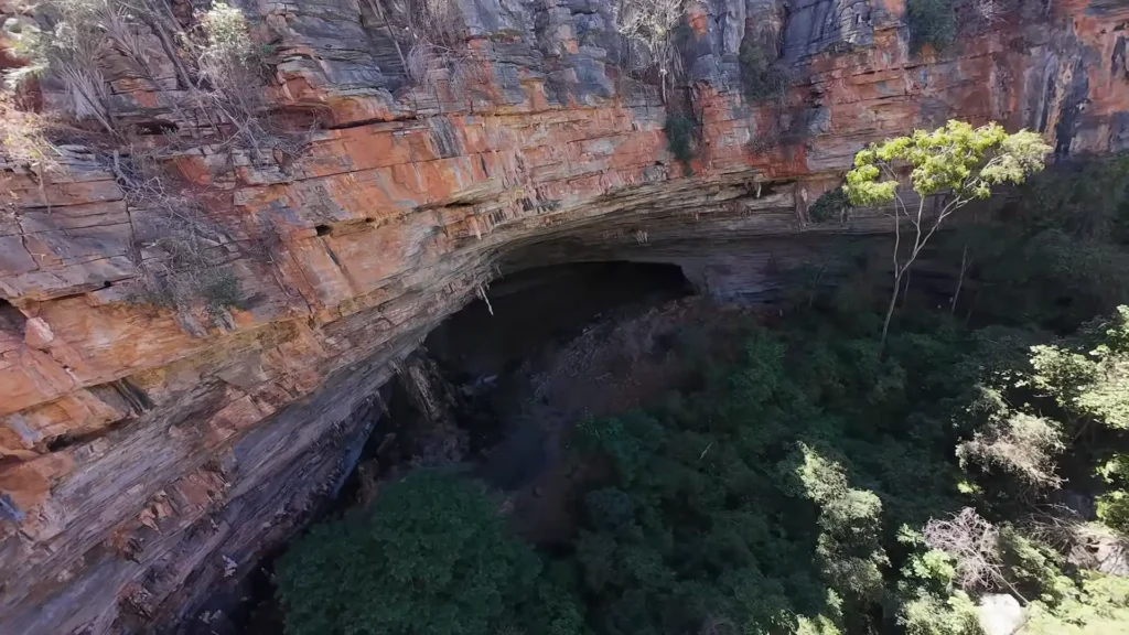 Entrada imensa de uma caverna vista de cima com paredões rochosos avermelhados.