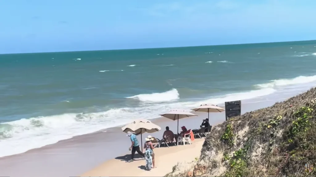 Vista panorâmica da praia com mar azul, ondas, areia clara e guarda-sóis bege em frente ao Coliseum Beach Resort no Ceará.