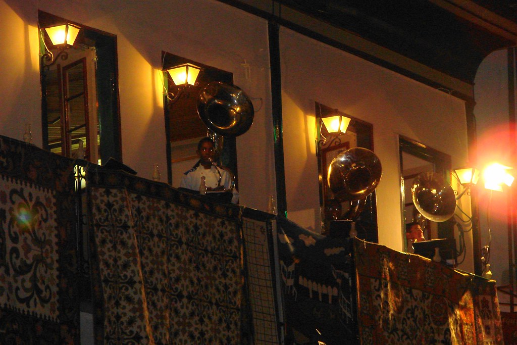 Músicos tocando instrumentos de sopro (sousafones) nas sacadas de um casarão colonial iluminado e decorado com tapetes estampados durante a noite na Vesperata de Diamantina.