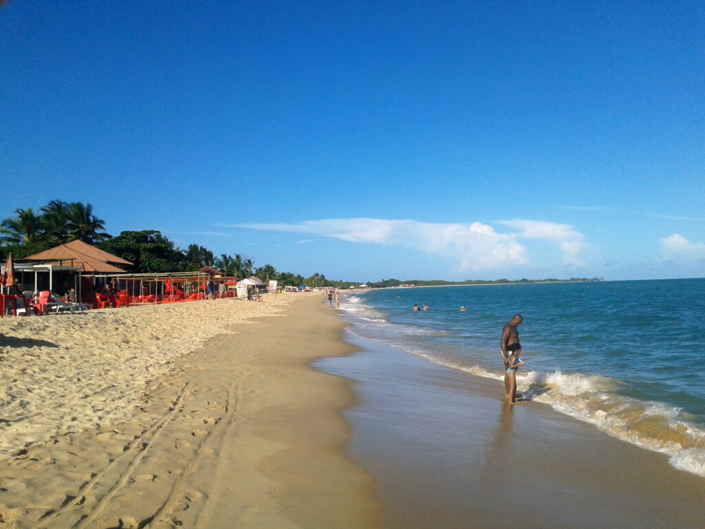 Família aproveitando o dia na areia da Praia de Taperapuã em Porto Seguro, com mar azul calmo e barracas de praia ao fundo.