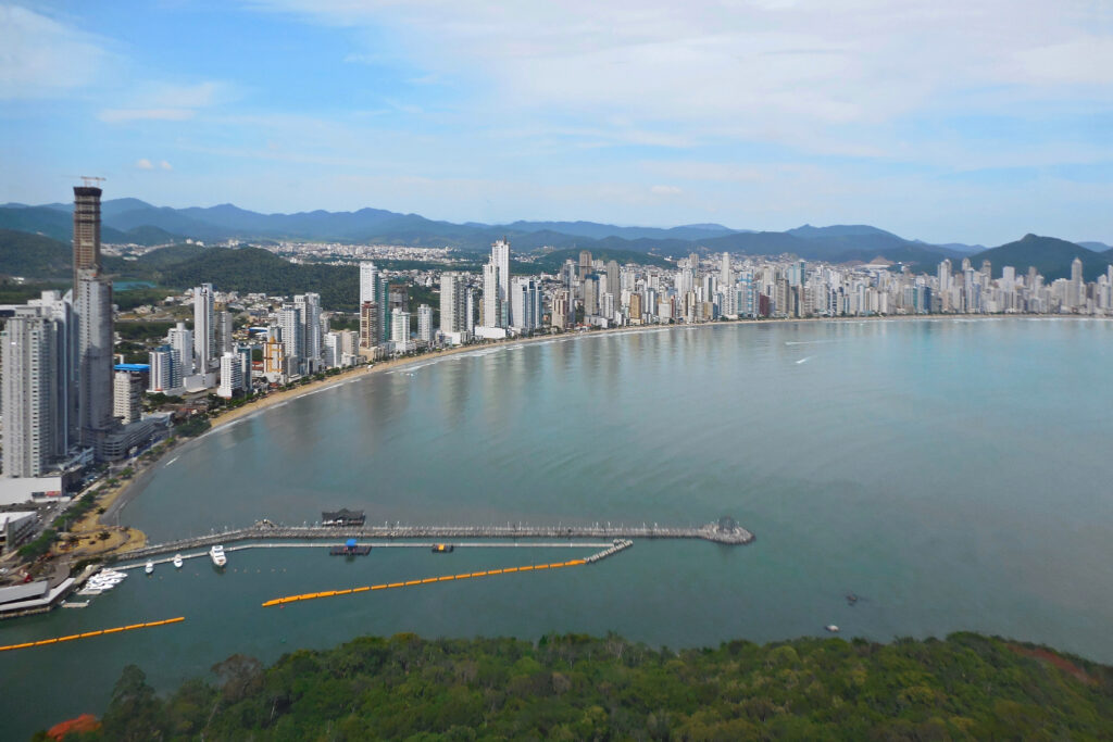 Vista aérea da orla de Balneário Camboriú com seus arranha-céus modernos, a faixa de areia curva e o mar calmo.