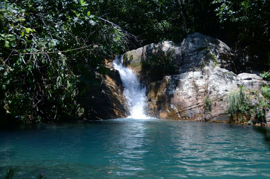 Pequena queda d'água desaguando em um poço de cor azul turquesa cercado por pedras e mata.