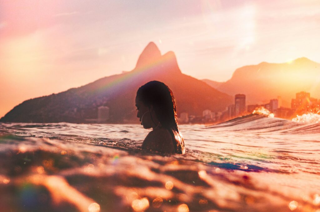 Mulher mergulhando no mar durante um pôr do sol dourado na Praia de Ipanema, com o Morro Dois Irmãos ao fundo.