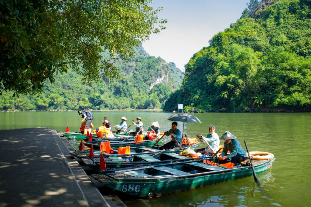 Vários barcos a remo verdes alinhados na margem de um rio com turistas embarcando.