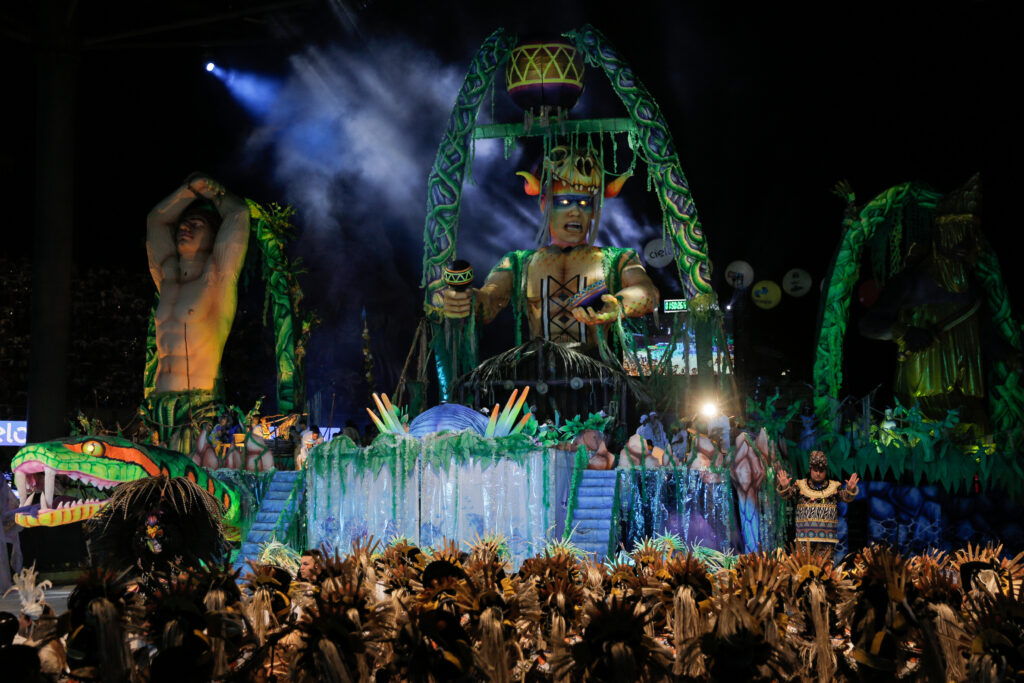 Carro alegórico iluminado à noite no Festival de Parintins, exibindo uma figura mitológica gigante cercada por folhagem e adereços, com centenas de dançarinos usando cocares de penas em primeiro plano.