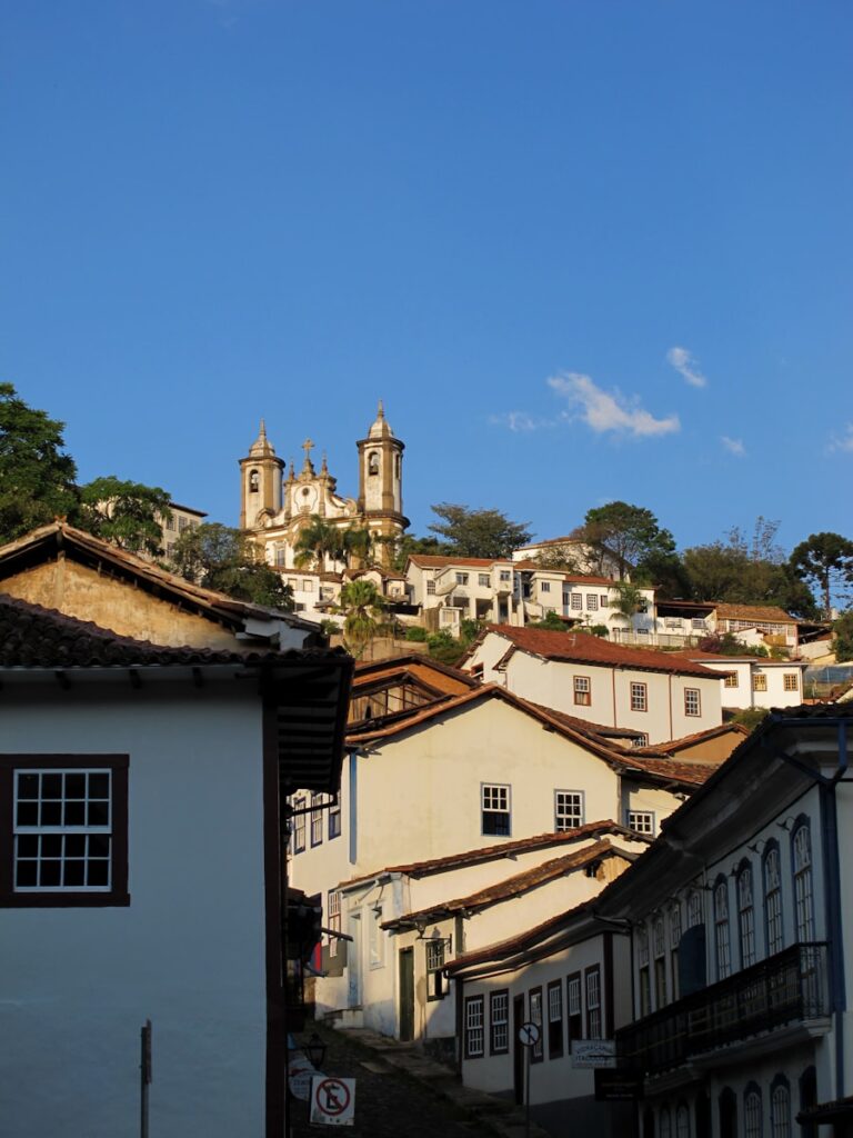 Ladeira de paralelepípedos cercada por casarões coloniais brancos, levando à Igreja de Santa Efigênia no topo do morro, sob um céu azul límpido.