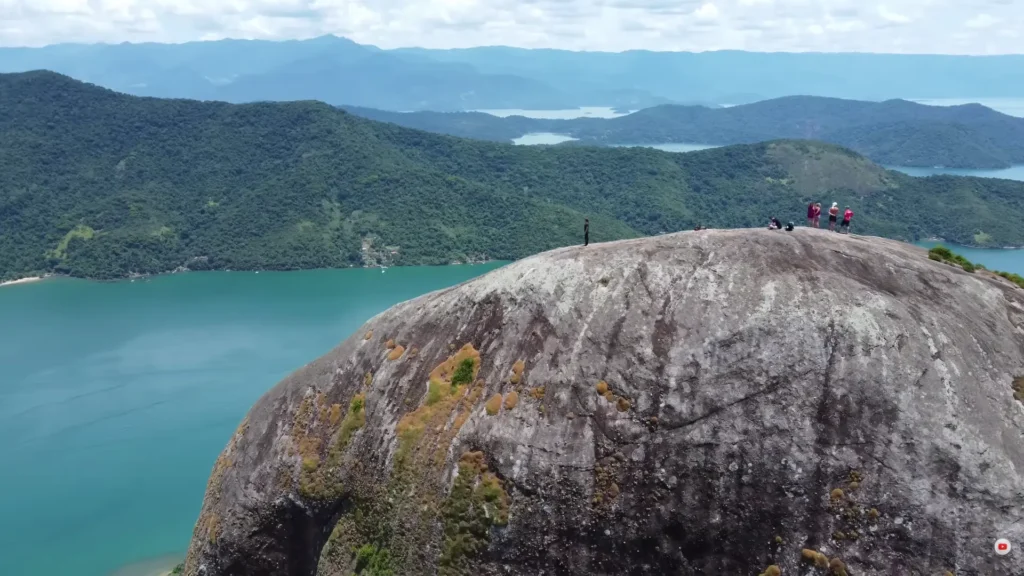 Turistas no topo da rocha do Pico do Pão de Açúcar do Mamanguá, observando a vista panorâmica do fiorde tropical e as águas verde-esmeralda.