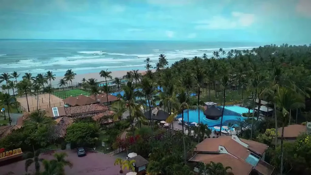 Vista aérea da costa mostrando as ondas do mar, a faixa de areia e o resort imerso em coqueiros.