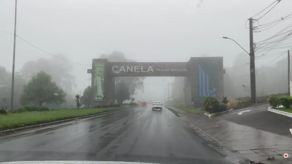 Pórtico de entrada da cidade de Canela envolto em forte neblina (cerração), com a frase "Canela Paixão Natural" visível, transmitindo clima de inverno.