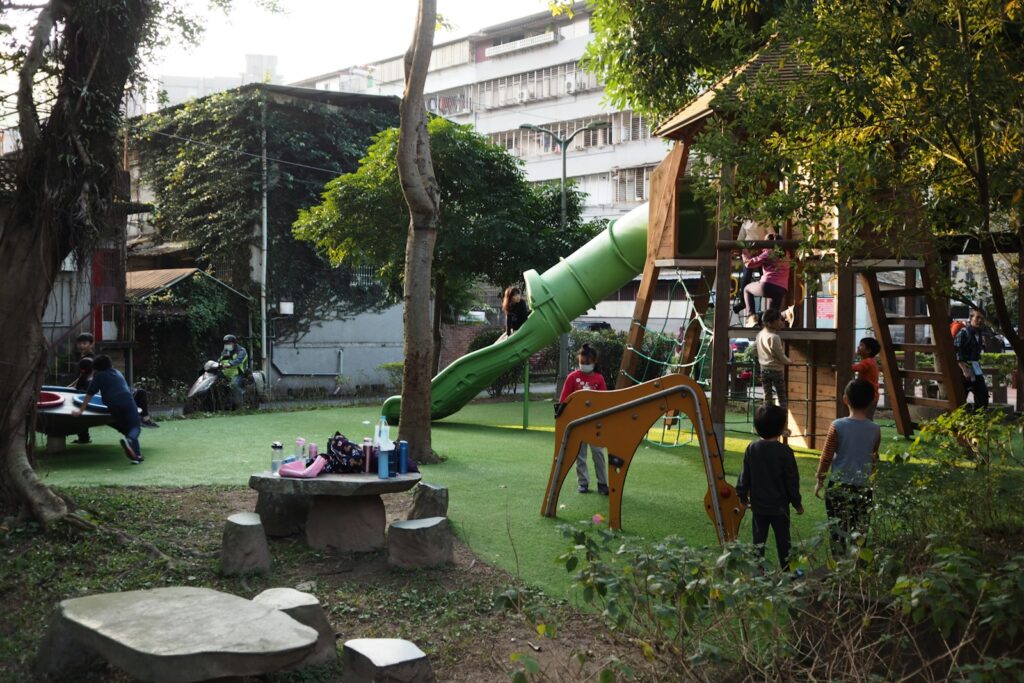 Crianças brincando em um playground ao ar livre com escorregador verde e casa de madeira cercada por árvores.
