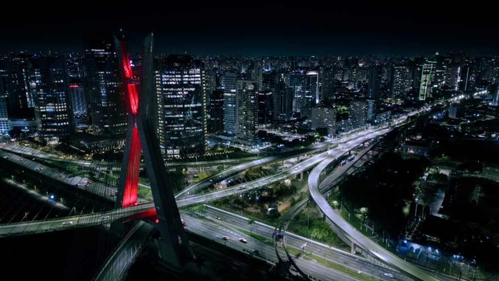  Vista aérea noturna da Ponte Estaiada em São Paulo, com seus cabos iluminados em vermelho vibrante e arranha-céus iluminados ao fundo.