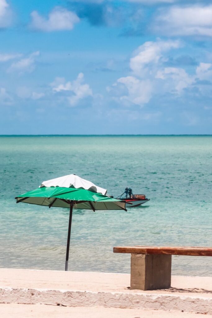 Peaceful beach view in Brazil with green umbrella, bench, and boat on turquoise ocean under blue sky.