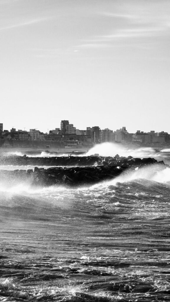 Black and white view of waves against the Mar del Plata coastline with cityscape in the background.