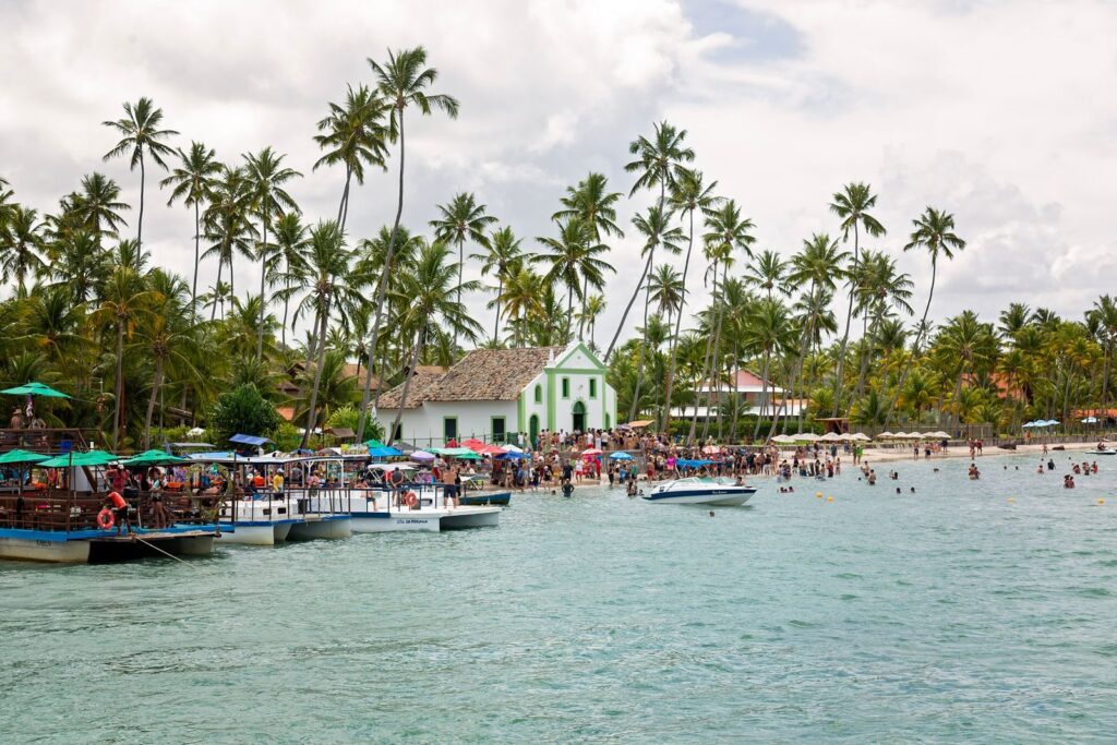Lively beach and boats near historic São Miguel dos Milagres church with palm backdrop.